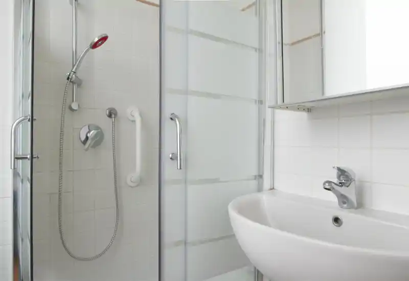 Bagno moderno e compatto con doccia vetro, soffione a mano, maniglione di sicurezza e lavabo bianco — piastrelle chiare e rubinetteria cromata.