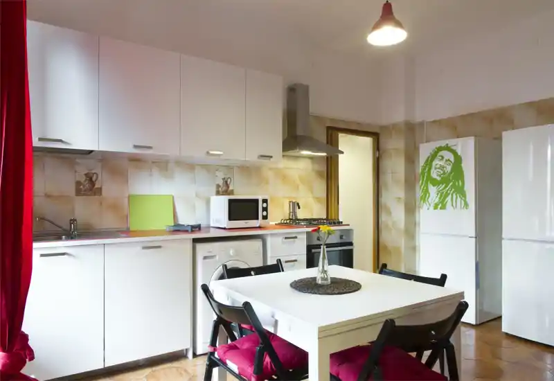 Bright, well-equipped kitchen with white cabinets, a dining table and red cushion chairs. The space includes a gas hob, microwave, washing machine and a fridge with decorative decal.
