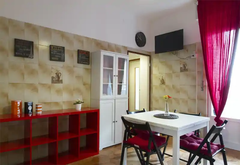 Bright, cozy dining area with a white table, four cushioned chairs and a large window dressed with red curtains; decorative shelving and a glass-front cabinet add storage and style.
