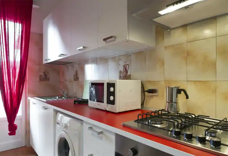Compact, modern kitchen with red countertop, gas hob, microwave and sink; warm tiled backsplash and a bright window with a red curtain.