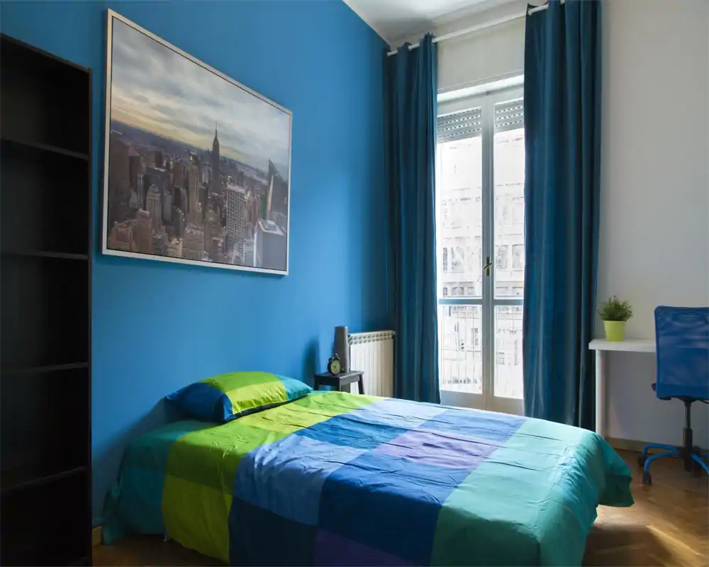 Camera da letto luminosa e moderna con letto matrimoniale, biancheria colorata, grande stampa della città su una parete blu e scrivania vicino alla finestra con luce naturale.