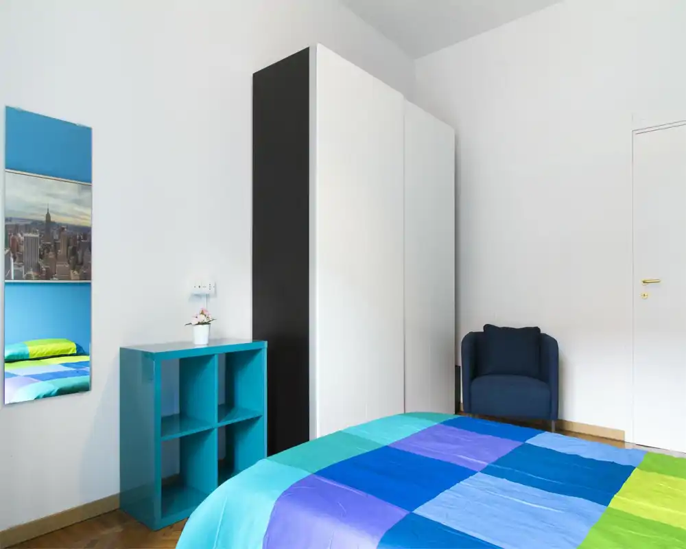 Camera da letto luminosa e moderna con copriletto colorato, grande armadio bianco e una poltrona blu scuro — stile semplice e pulito con ottima luce naturale.