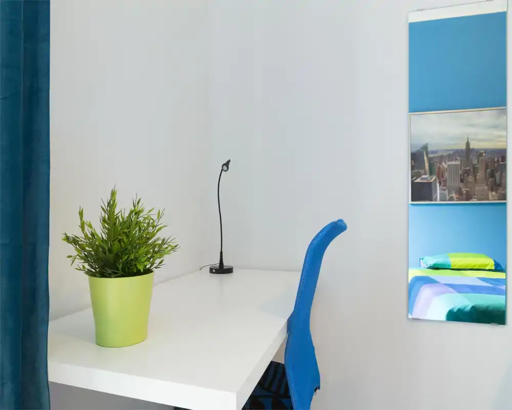 Angolo della camera luminoso e minimal con scrivania bianca e sedia blu; il letto è visibile solo come riflesso nello specchio, mentre la postazione e la pianta decorativa sono in primo piano.