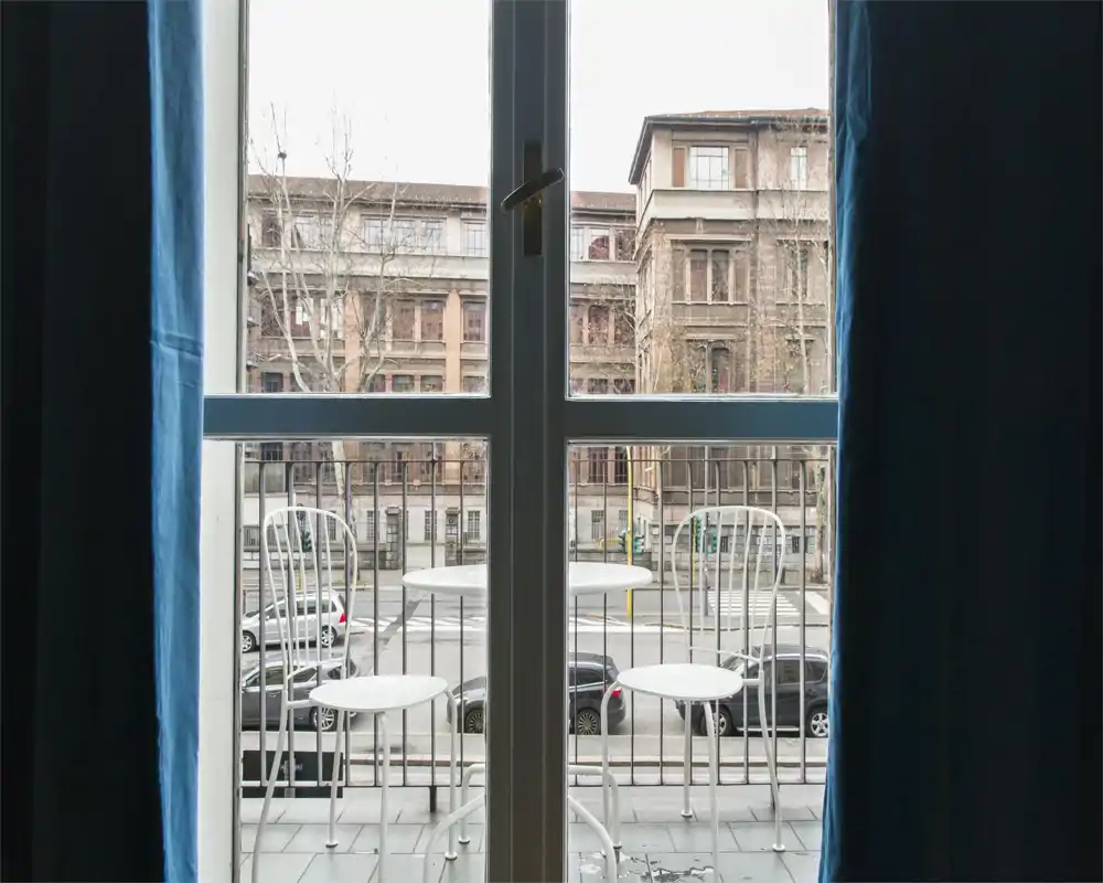 Piccolo balcone privato con due sedie in metallo e un tavolino rotondo, visto dall'interno attraverso una porta finestra; strada urbana e edifici visibili oltre.