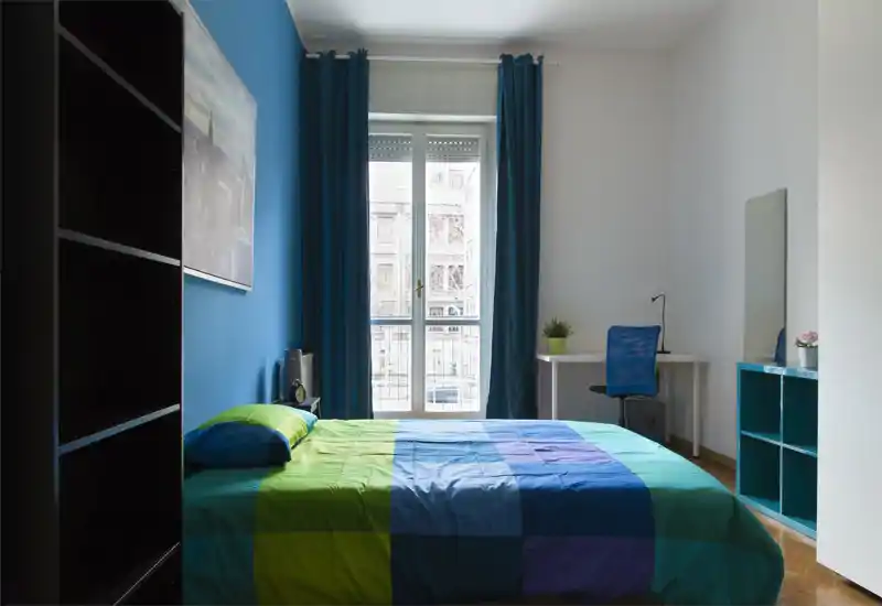 Chambre moderne et lumineuse avec un grand lit double au premier plan, mur bleu en accent et fenêtre pleine hauteur apportant une belle lumière naturelle. Un bureau compact, des étagères et des plantes décoratives complètent l'agencement fonctionnel.