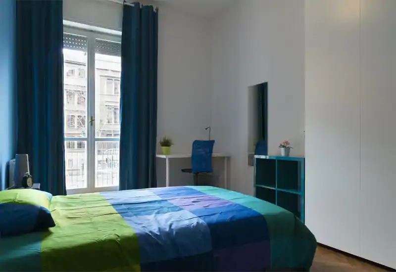Chambre moderne et lumineuse avec un grand lit aux couettes colorées, une fenêtre avec rideaux foncés, un petit bureau et des décorations — propre et confortable.