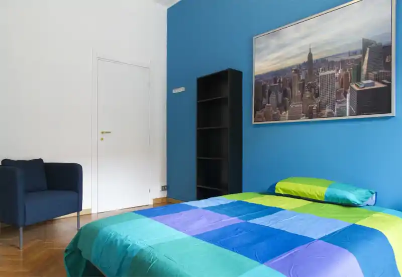 Chambre moderne et lumineuse avec lit coloré, mur bleu et grand tableau urbain. Un fauteuil et une étagère complètent l'agencement soigné et contemporain.
