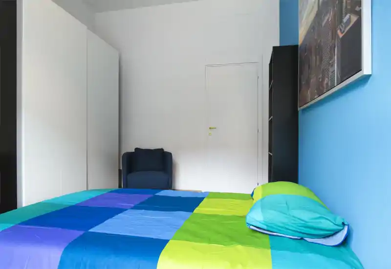 Chambre lumineuse et moderne avec un grand lit coloré au premier plan, un mur d'accent bleu et un petit fauteuil près d'une armoire blanche.