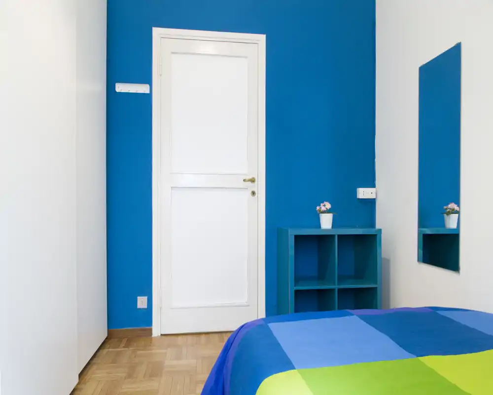 Chambre lumineuse et soignée avec une couverture colorée, un mur bleu et une petite étagère décorée de plantes — idéale pour une annonce accueillante.