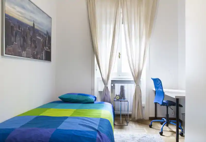 Chambre simple lumineuse et bien rangée avec une couette colorée, une table de chevet et un coin bureau près de la fenêtre — parfaite pour un étudiant ou une personne seule.