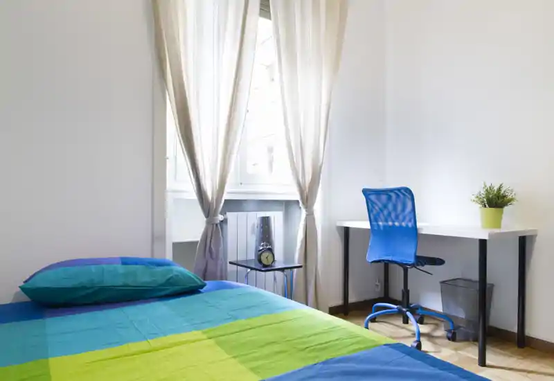 Chambre lumineuse et moderne avec un lit coloré, des rideaux légers et un petit espace de travail équipé d'une chaise ergonomique bleue — idéale pour se reposer et télétravailler.