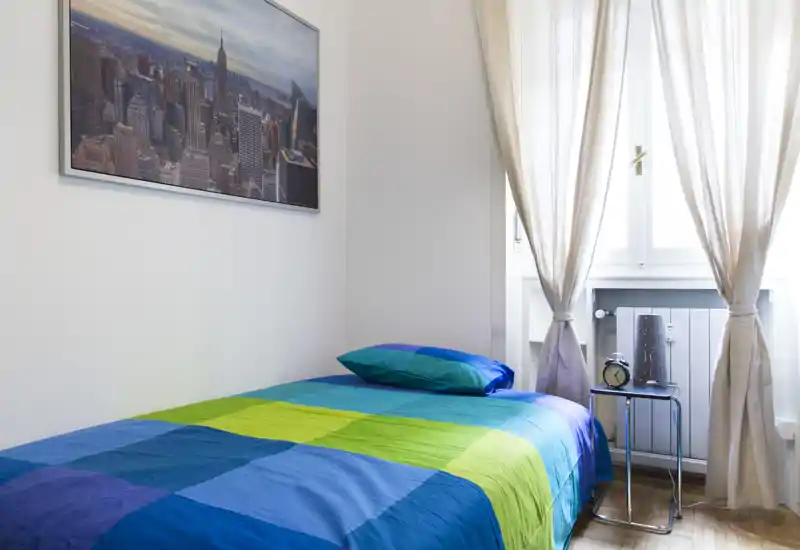 Chambre simple et lumineuse avec un lit coloré, une table de chevet avec réveil et une belle lumière naturelle provenant de la fenêtre aux rideaux clairs.