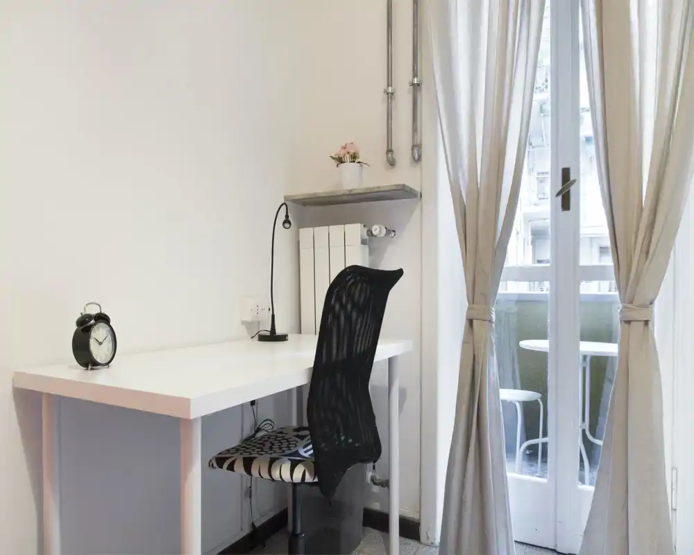 Angolo luminoso e minimal della camera adibito a postazione di lavoro con scrivania bianca, sedia ergonomica in rete, lampada da tavolo e tende che si aprono su un balcone.