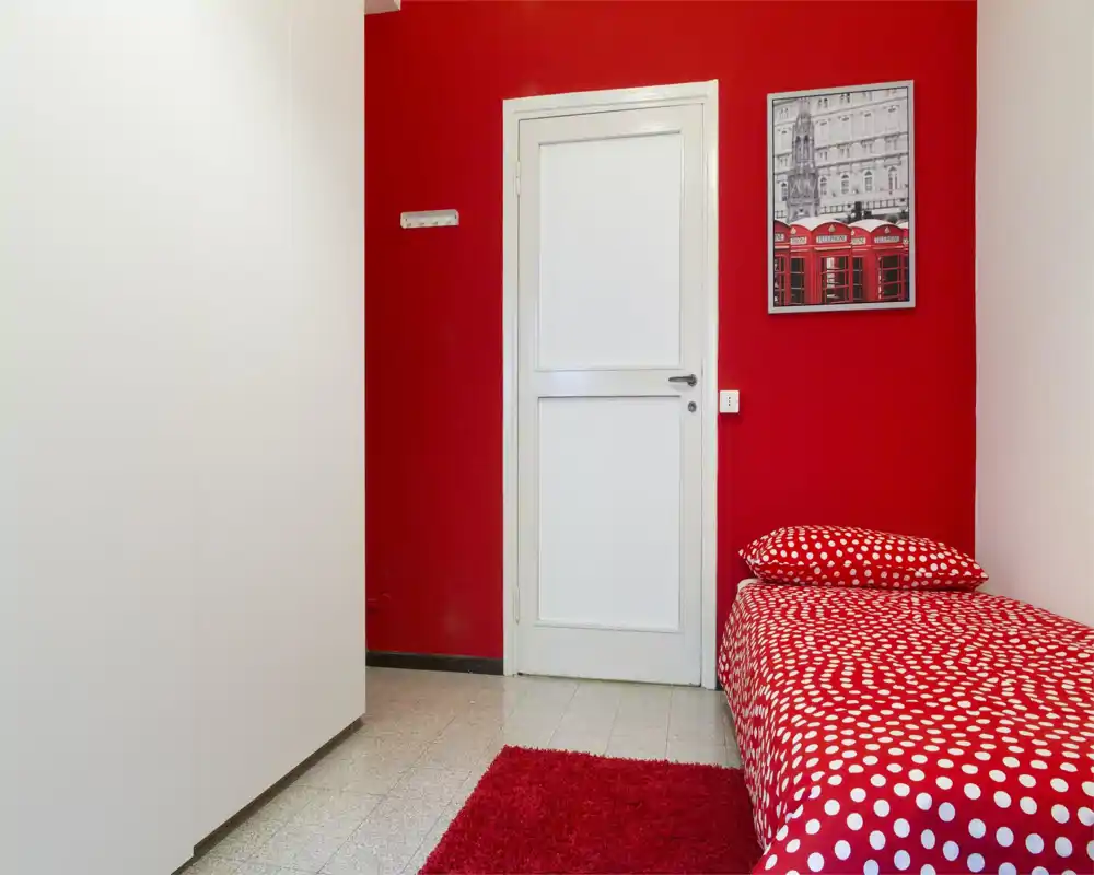 Camera singola compatta con parete rossa, biancheria a pois e piccolo tappeto; ordinata e luminosa, ideale per una persona.