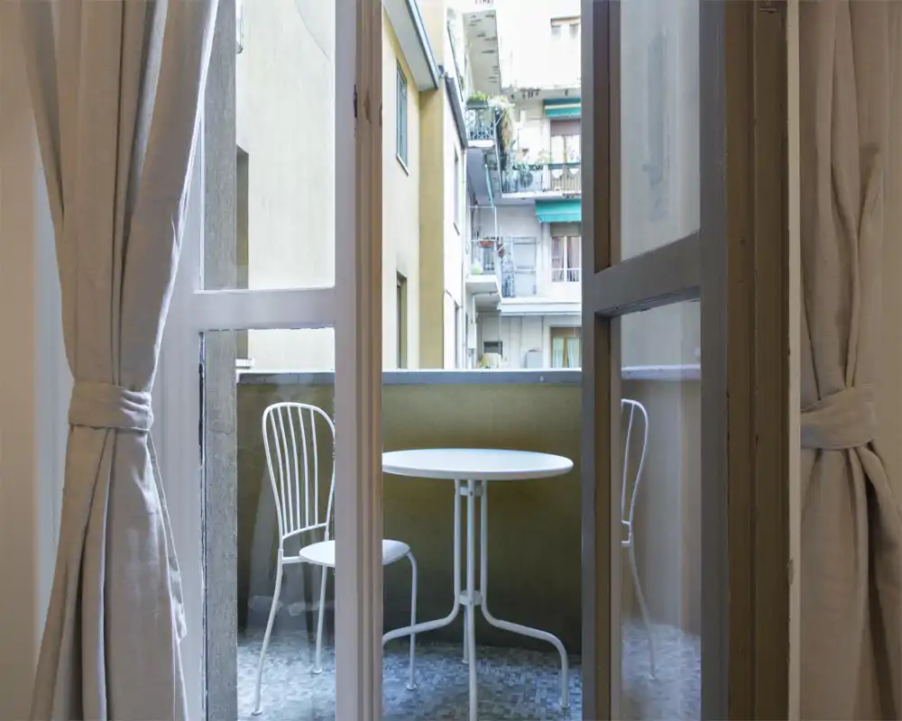 Piccolo balcone privato con tavolino rotondo e due sedie in metallo, visibile attraverso le tende legate; perfetto per il caffè mattutino con vista sugli edifici vicini.