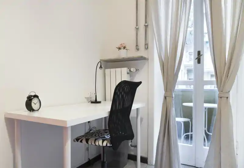 Angolo luminoso e minimal della camera adibito a postazione di lavoro con scrivania bianca, sedia ergonomica in rete, lampada da tavolo e tende che si aprono su un balcone.