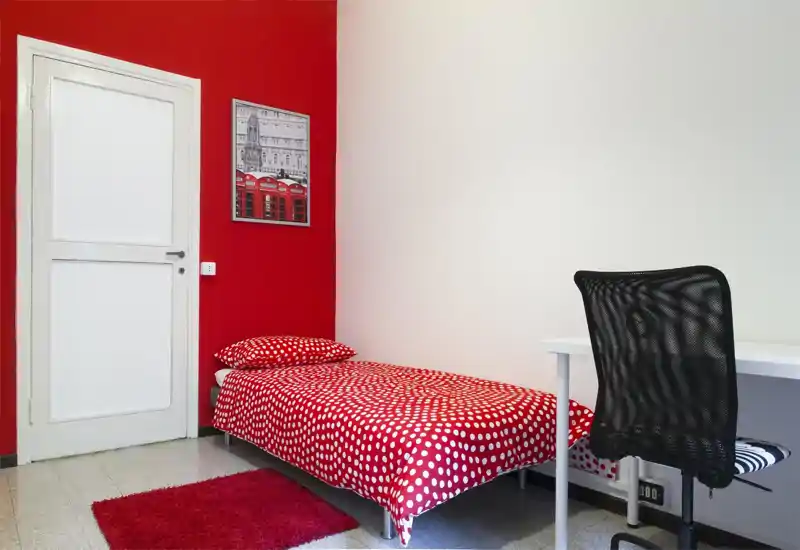 Camera da letto compatta e luminosa con letto singolo e copriletto rosso a pois, tappeto coordinato e una piccola scrivania bianca con sedia ergonomica — perfetta per studenti o viaggiatori singoli.