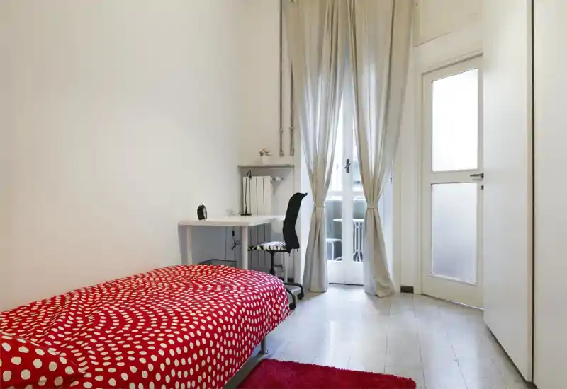 Camera singola luminosa e minimale con copriletto rosso a pois, scrivania e luce naturale dalla porta balcone — ideale per uno studente o single.
