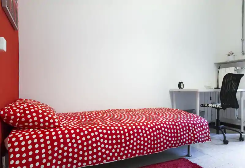 Camera da letto singola luminosa e ordinata con copriletto rosso a pois e una scrivania semplice con sedia — ideale per uno studente o una persona sola.