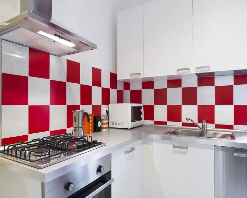Cucina compatta e moderna con piano cottura a gas in acciaio, forno incorporato, lavello e vivace paraspruzzi a scacchi rosso e bianco — luminosa, pulita e funzionale.