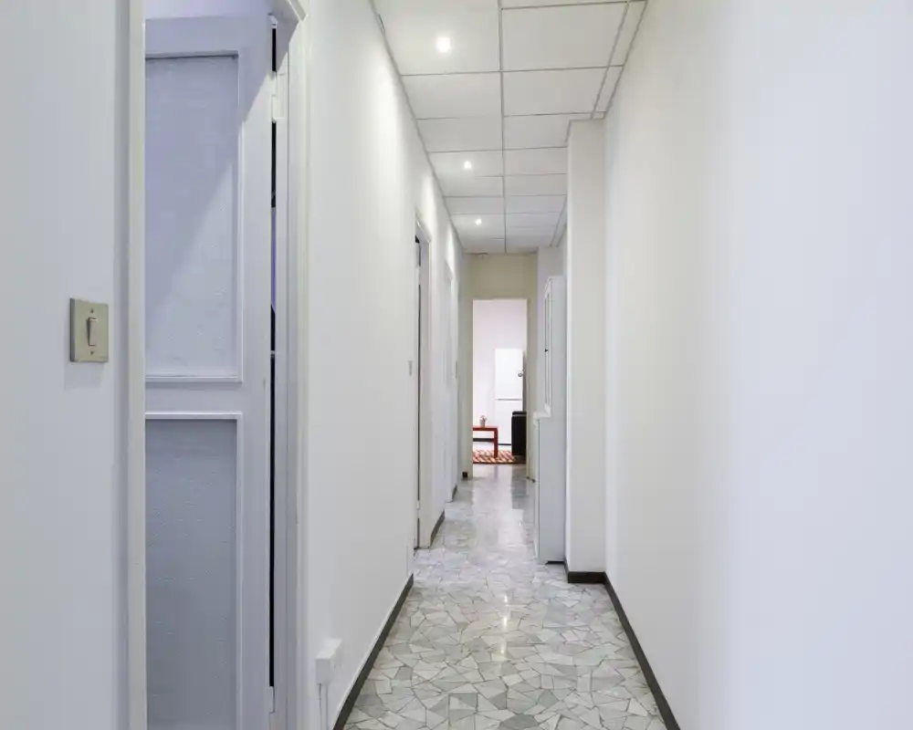 Corridoio lungo e pulito con pavimento a mosaico, pareti bianche e faretti a soffitto che conducono ad altre stanze; arredo neutro e minimale adatto a una proprietà in affitto.