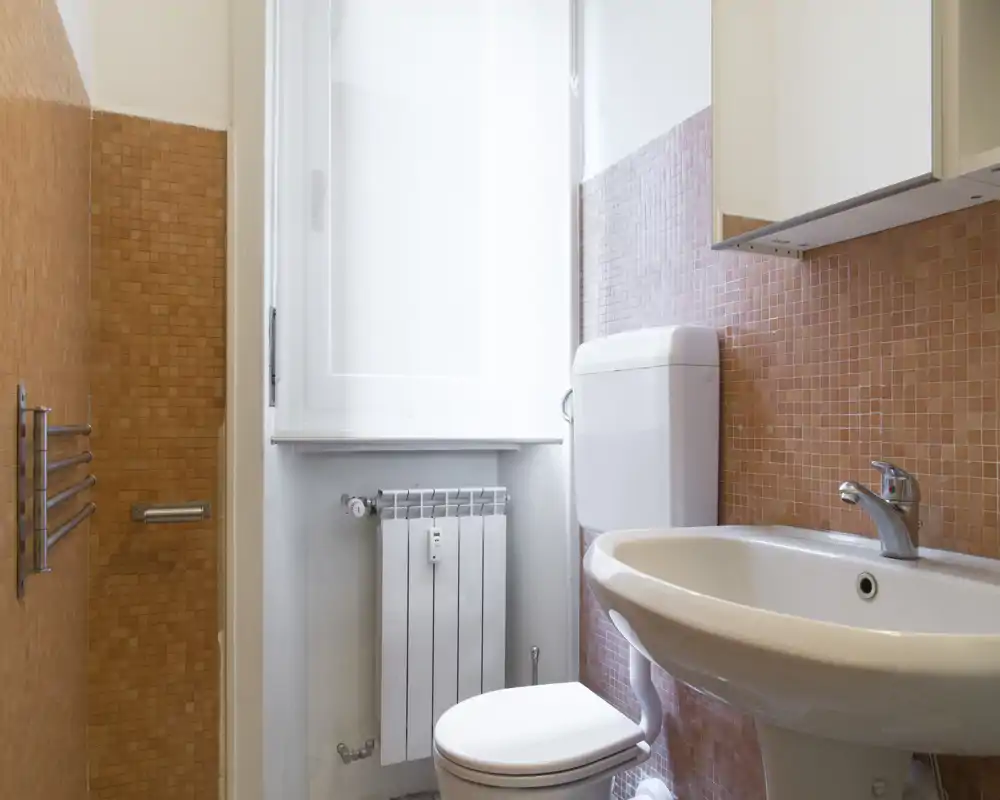 Bagno compatto e luminoso con lavabo bianco, WC e piastrelle mosaico arancioni; una finestra illumina l'ambiente e un pensile a muro offre spazio di servizio.