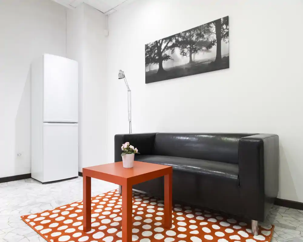 Piccolo soggiorno moderno con divano in pelle nera, lampada da terra sottile, tavolino arancione e tappeto a fantasia, pareti bianche e illuminazione luminosa.