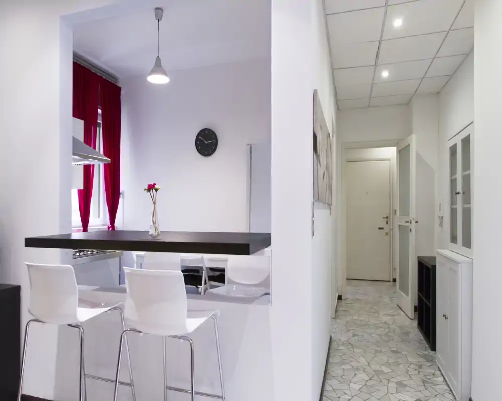 Angolo cottura open-space con piano rialzato a colazione, sgabelli bianchi moderni e top scuro. Interno luminoso con tende rosse e lampada a sospensione, perfetto per appartamenti compatti in città.