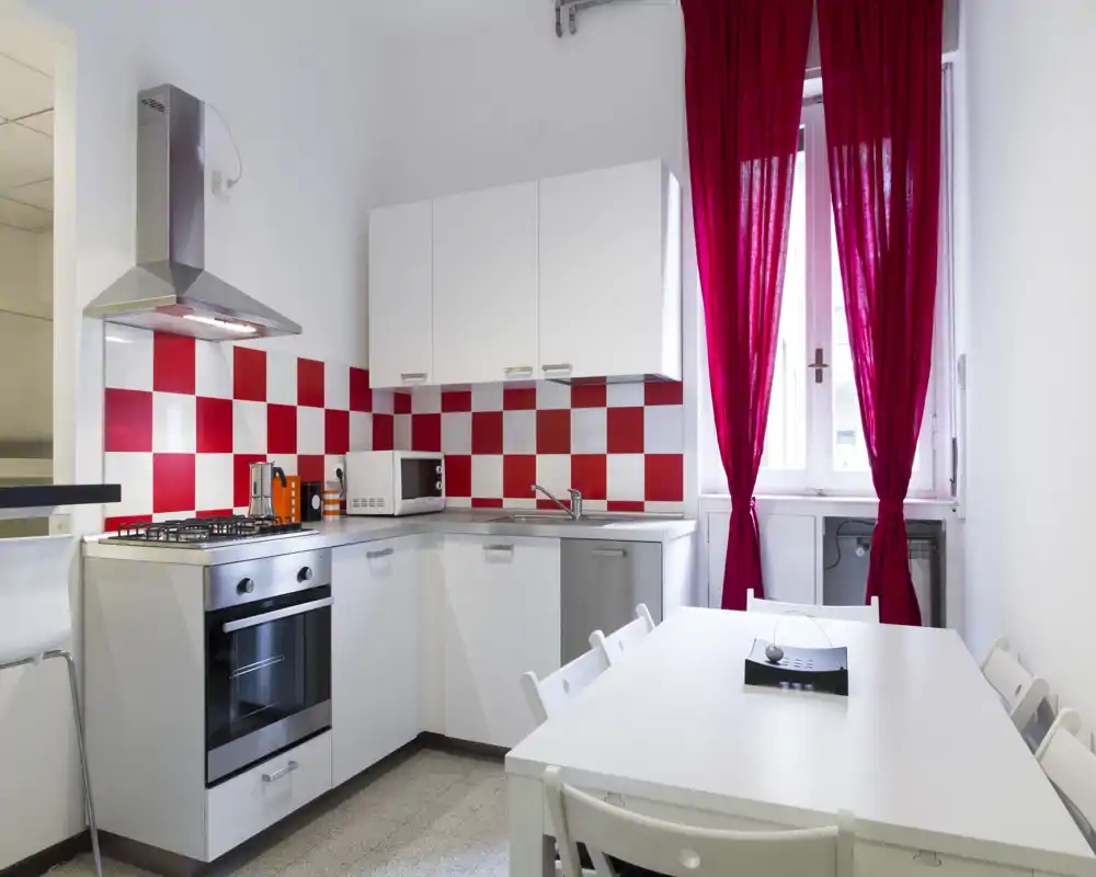 Cucina compatta e luminosa con mobili bianchi, paraschizzi a quadretti rosso e bianco, forno e cappa in acciaio e un piccolo tavolo da pranzo vicino alla finestra con tende rosse.