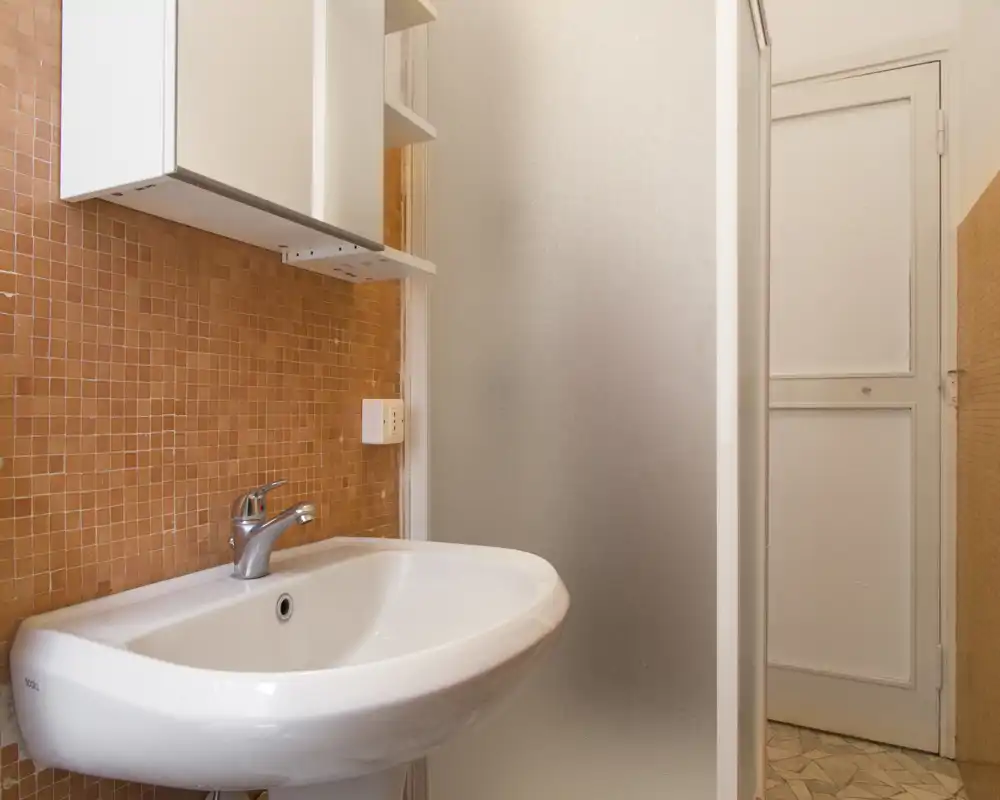 Piccola area bagno con lavabo in ceramica bianca, miscelatore monocomando, rivestimento a mosaico arancione e parete divisoria in vetro satinato; ambiente pulito e illuminato in modo semplice.