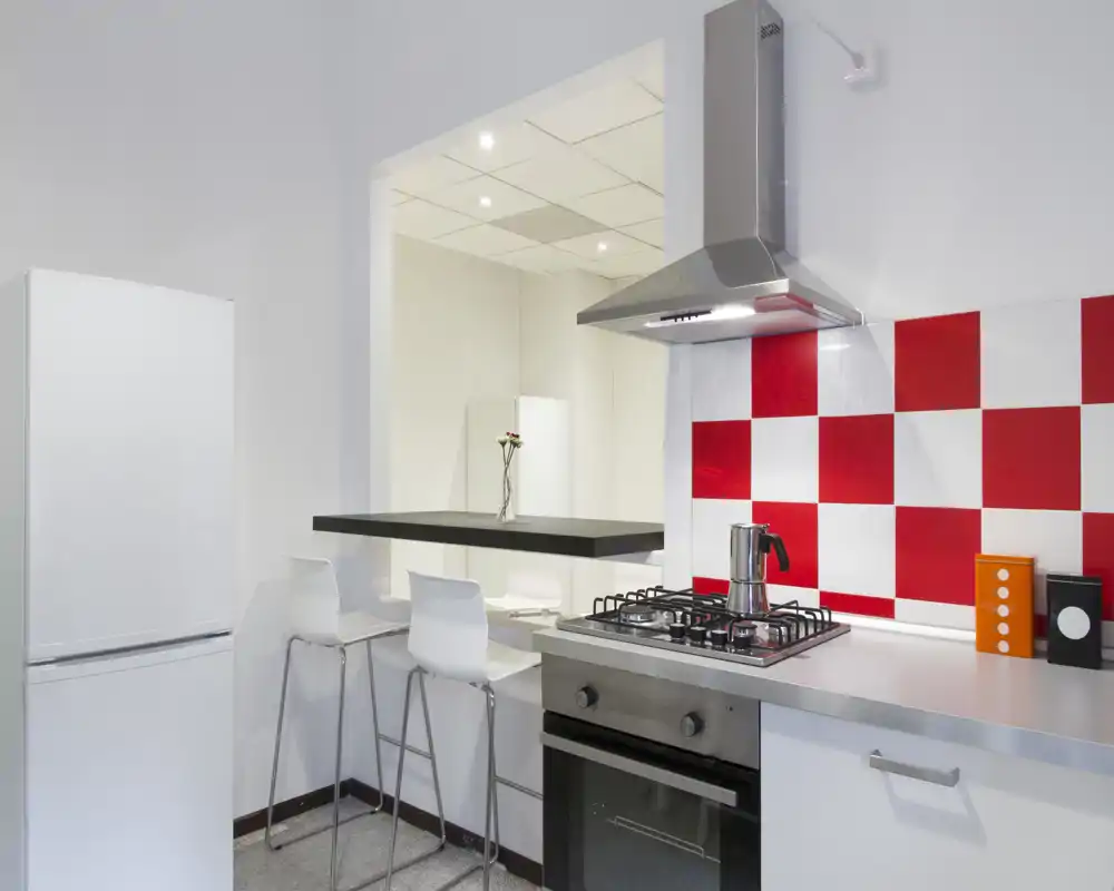 Cucina moderna e compatta con forno in acciaio, piano cottura a gas e paraschizzi a quadri rossi e bianchi. Bancone con sgabelli e frigorifero rendono lo spazio pratico e luminoso.