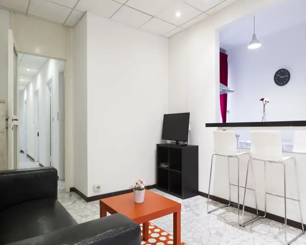 Area giorno moderna e luminosa con divano nero, mobile TV compatto e un piccolo tavolino da caffè arancione; passaggio aperto verso la cucina con sgabelli al banco.