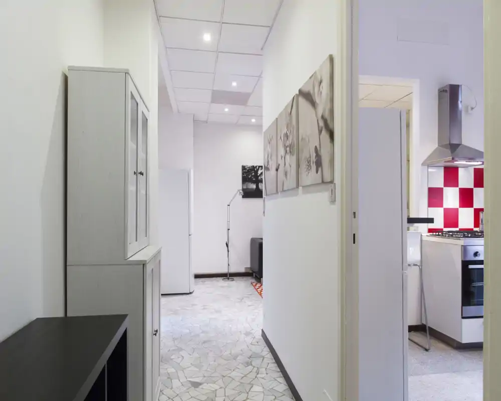 Vista di un corridoio luminoso con la cucina parzialmente visibile attraverso una porta aperta; la cucina ha un paraschizzi a scacchi rosso e bianco, cappa in acciaio e elettrodomestici moderni.