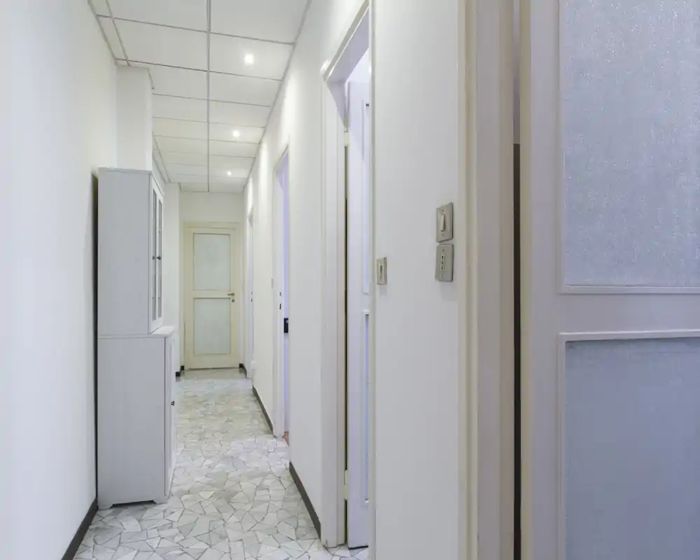Corridoio d'ingresso luminoso e stretto con pavimento a mosaico, faretti a soffitto e pareti bianche; presente un mobile contenitore e diverse porte lungo il corridoio.