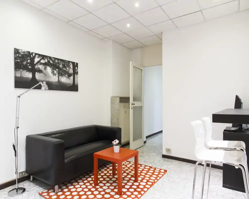 Soggiorno ben illuminato con divano nero per due persone, un piccolo tavolino arancione su un tappeto a pois e una lampada da terra; arredo minimalista e pareti neutre per un ambiente moderno e luminoso.