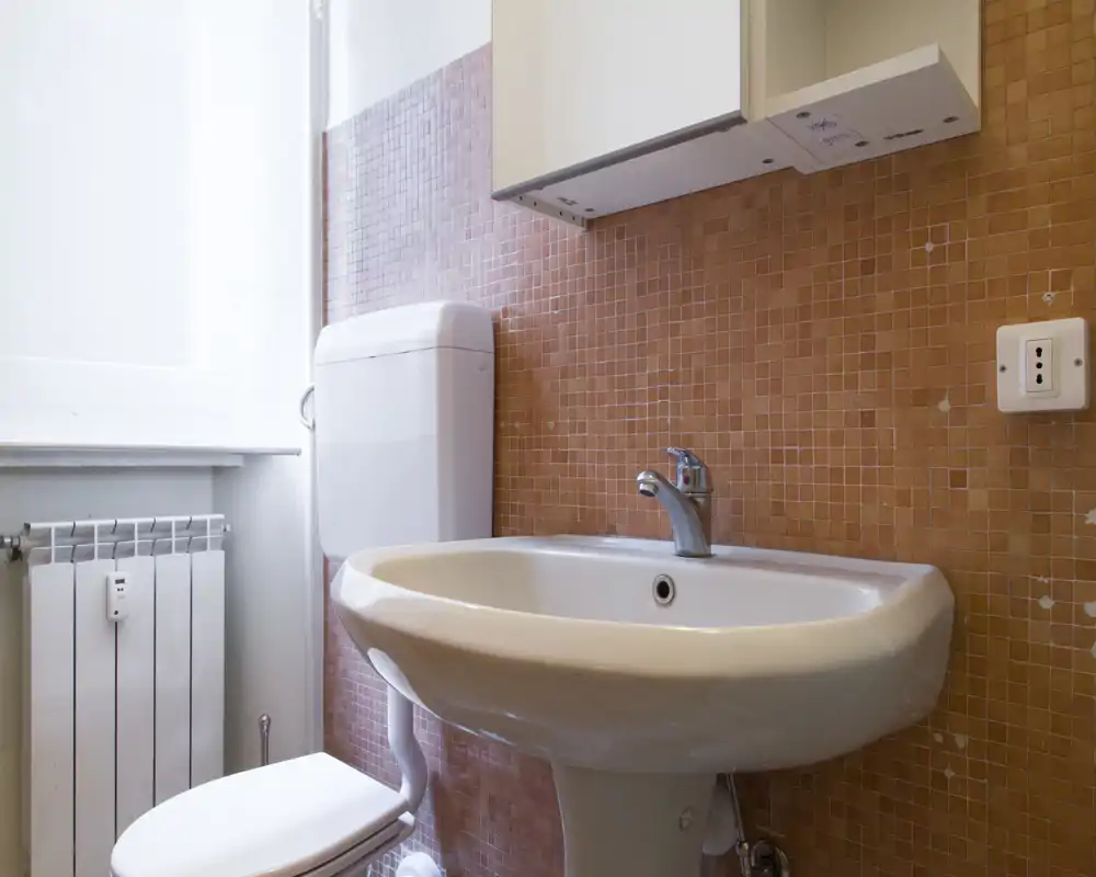 Bagno compatto con lavabo a colonna bianco, rubinetto cromato e WC davanti a piastrelle mosaico arancioni, illuminato da luce naturale proveniente dalla finestra.