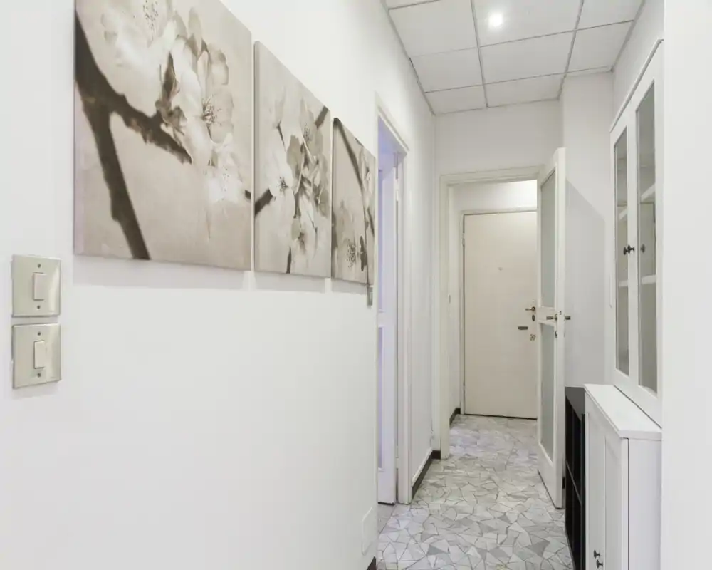 Ingresso luminoso e minimal con pavimento piastrellato, pareti bianche e un trittico decorativo a sinistra. L'immagine mostra la porta d'ingresso e un mobile contenitore, offrendo una chiara visione del corridoio.
