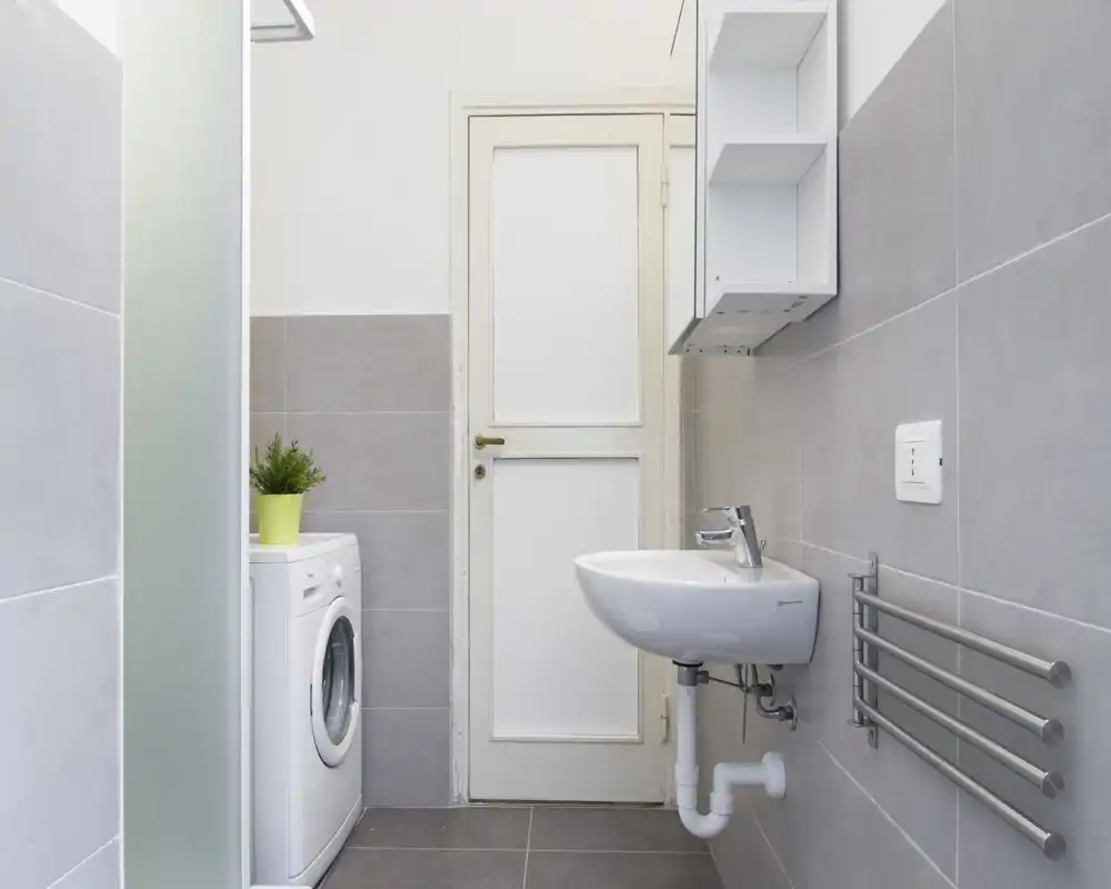Bagno pulito e luminoso con lavabo a parete, termoarredo e lavatrice accanto alla porta — piastrelle neutre moderne e arredi semplici e funzionali.