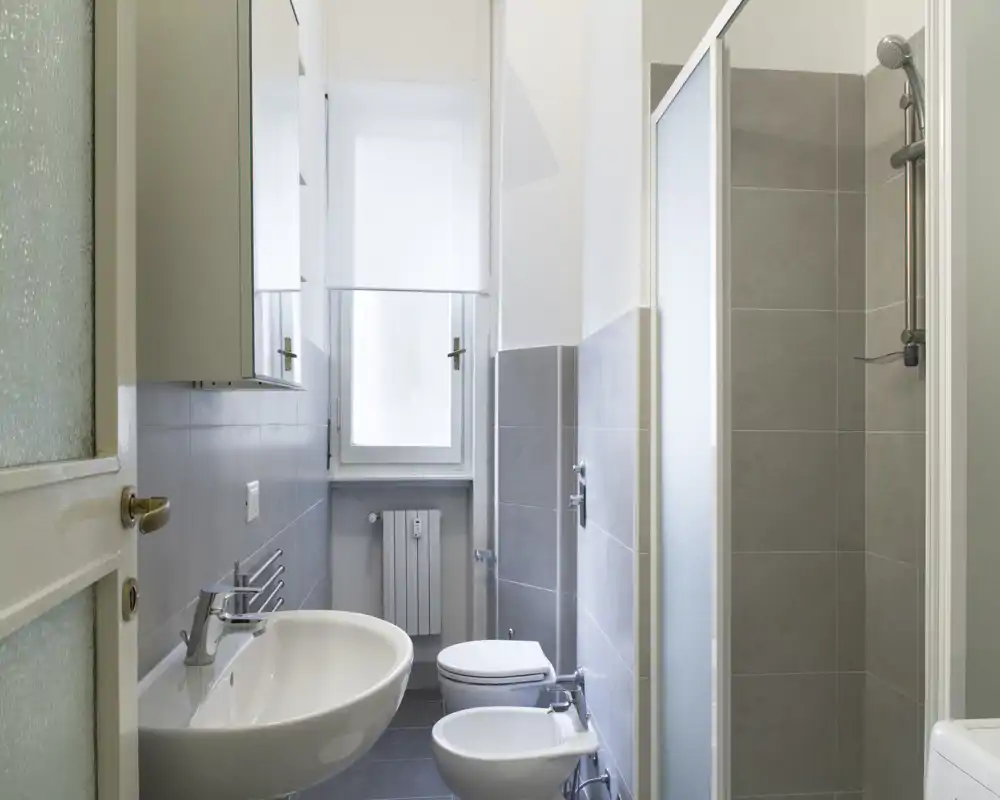 Bagno moderno e luminoso con lavabo a parete, bidet, wc e box doccia; piastrelle grigie neutre e finestra che garantisce luce naturale.