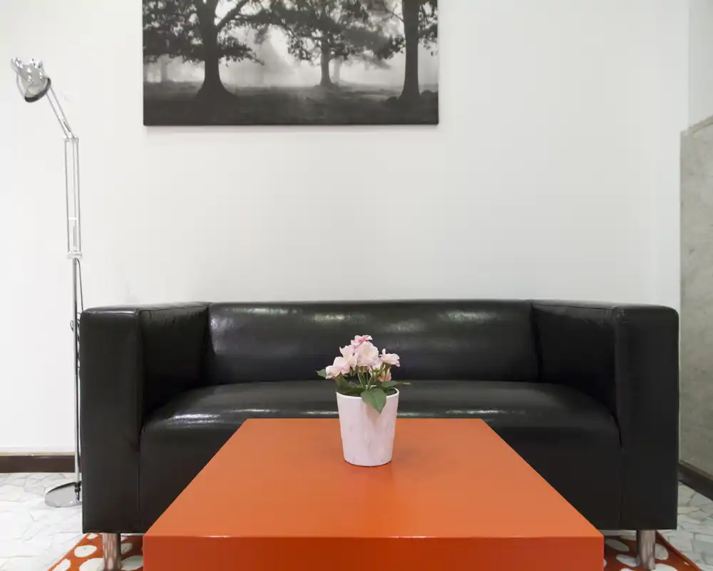 Soggiorno moderno con divano in pelle nero, tavolino da caffè arancione vivace e piccolo vaso di fiori — ambiente pulito, luminoso e accogliente.