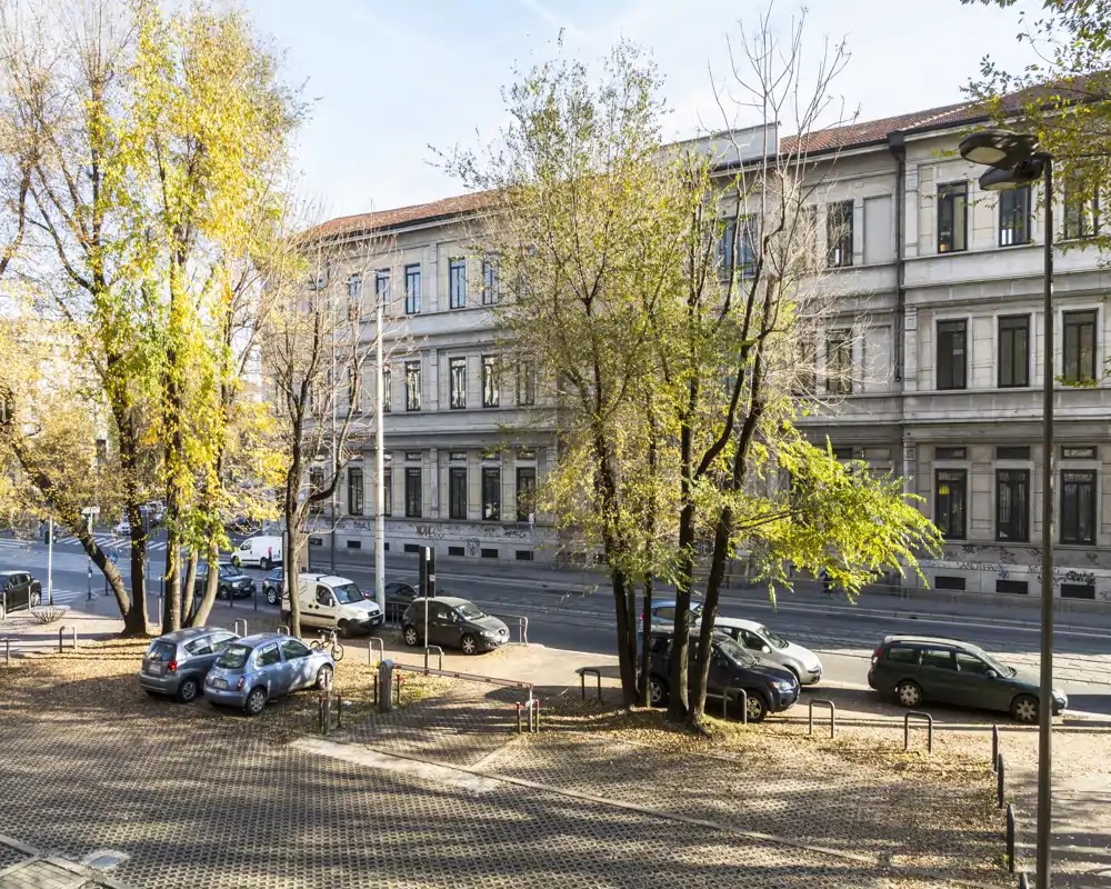 Vista esterna su strada con auto parcheggiate, alberi maturi e un edificio dall'aspetto storico di fronte; luce diurna nitida perfetta per mostrare il quartiere e la disponibilità di parcheggio sulla strada.