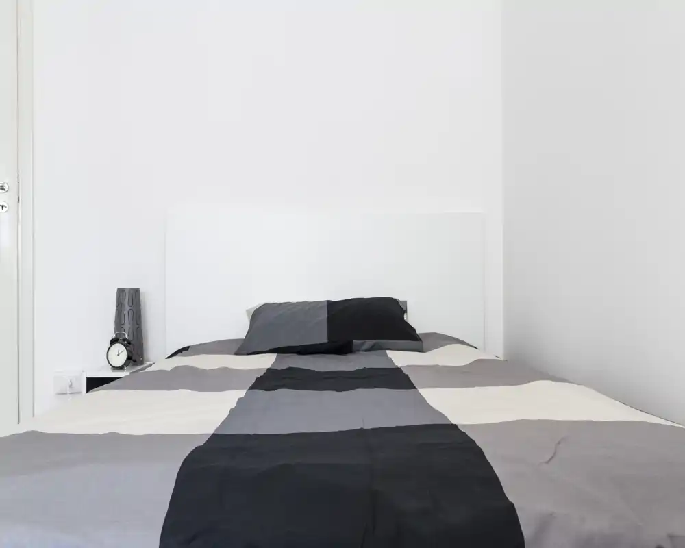 Camera da letto minimalista e luminosa con letto matrimoniale al centro, biancheria monocromatica e comodino con lampada e orologio.