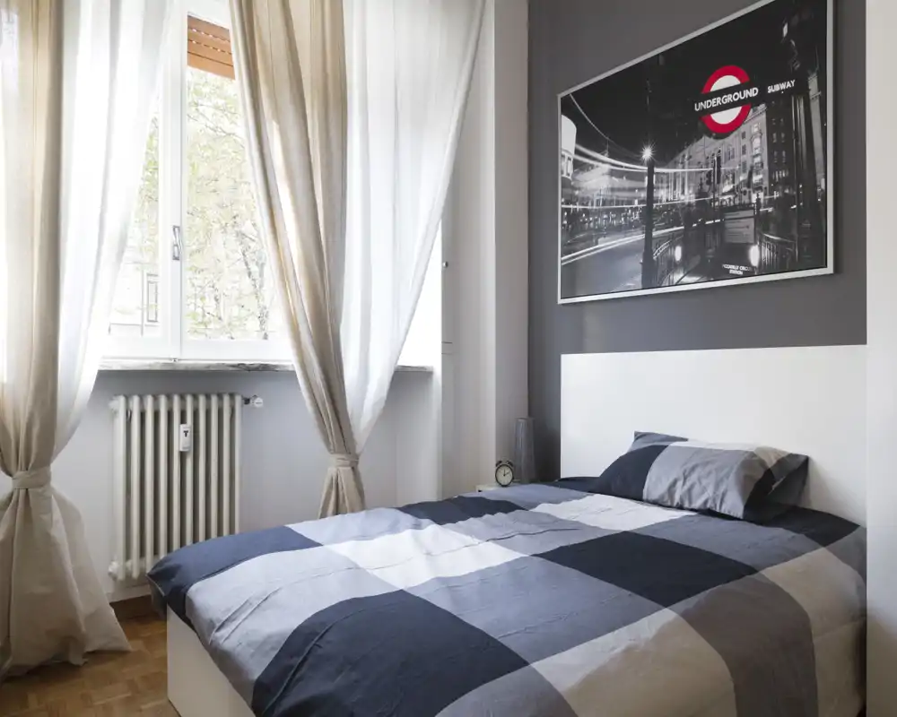 Camera da letto luminosa e di stile con letto matrimoniale, copriletto quadrettato grigio-blu, tende morbide e stampa cittadina sopra la testiera. La luce naturale riempie la stanza e i dettagli ordinati accanto al letto creano un’atmosfera accogliente e moderna.