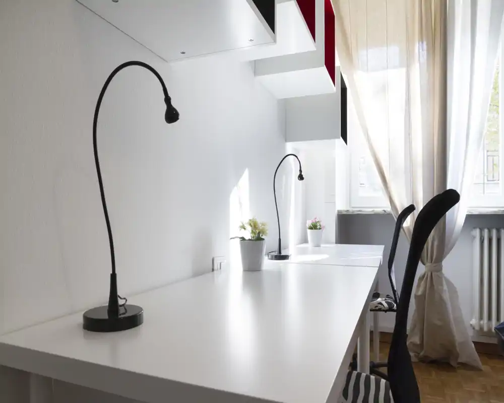 Spaziosa area di lavoro luminosa e minimal con una lunga scrivania bianca, due lampade da tavolo flessibili, sedie ergonomiche e piante vicino a una finestra con tende leggere.