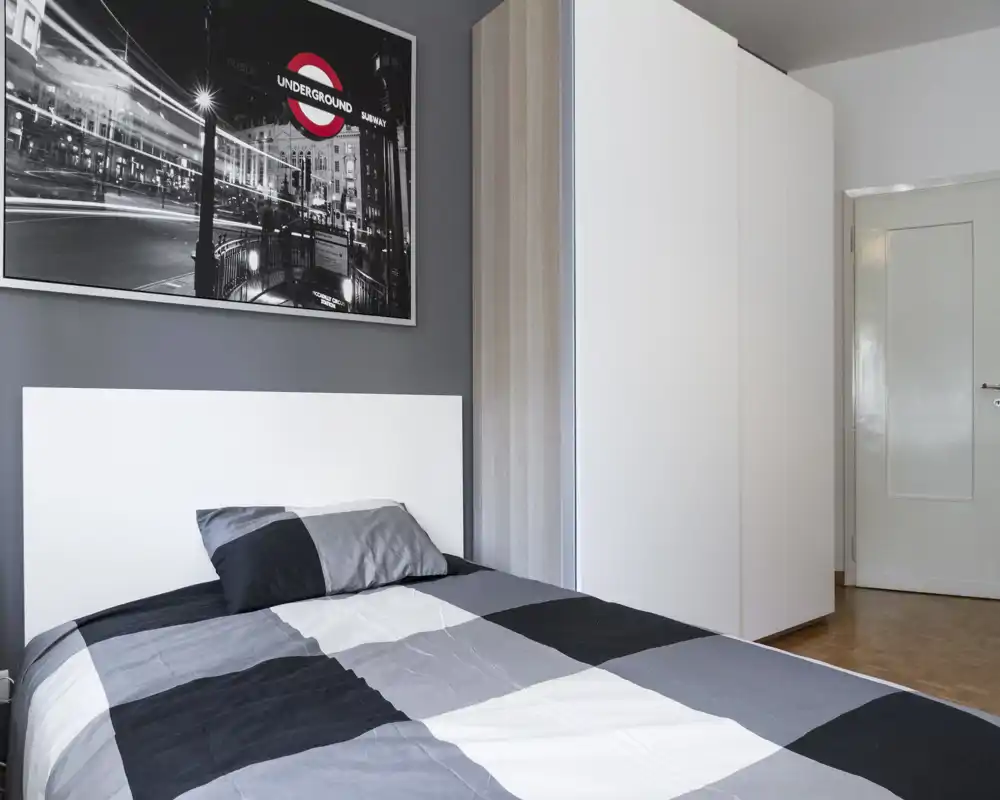 Camera da letto moderna e ben illuminata con letto matrimoniale con biancheria grigia e nera, testiera bianca e grande armadio. Palette neutra e quadro alle pareti creano un'atmosfera contemporanea e tranquilla, ideale per affitti brevi.