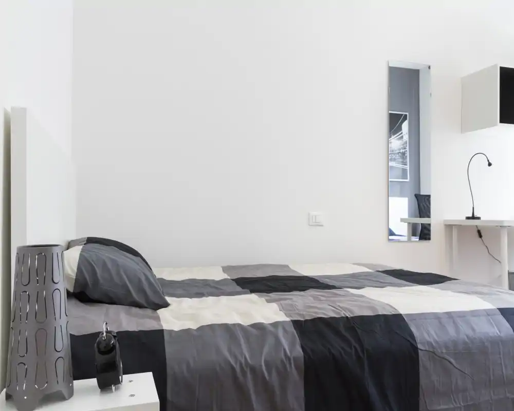 Camera da letto minimalista con un ampio letto matrimoniale con copriletto grigio e nero a quadri, una lampada da comodino e una piccola scrivania con specchio a parete. Luminoso e pulito, perfetto per annunci che puntano sulla semplicità moderna.