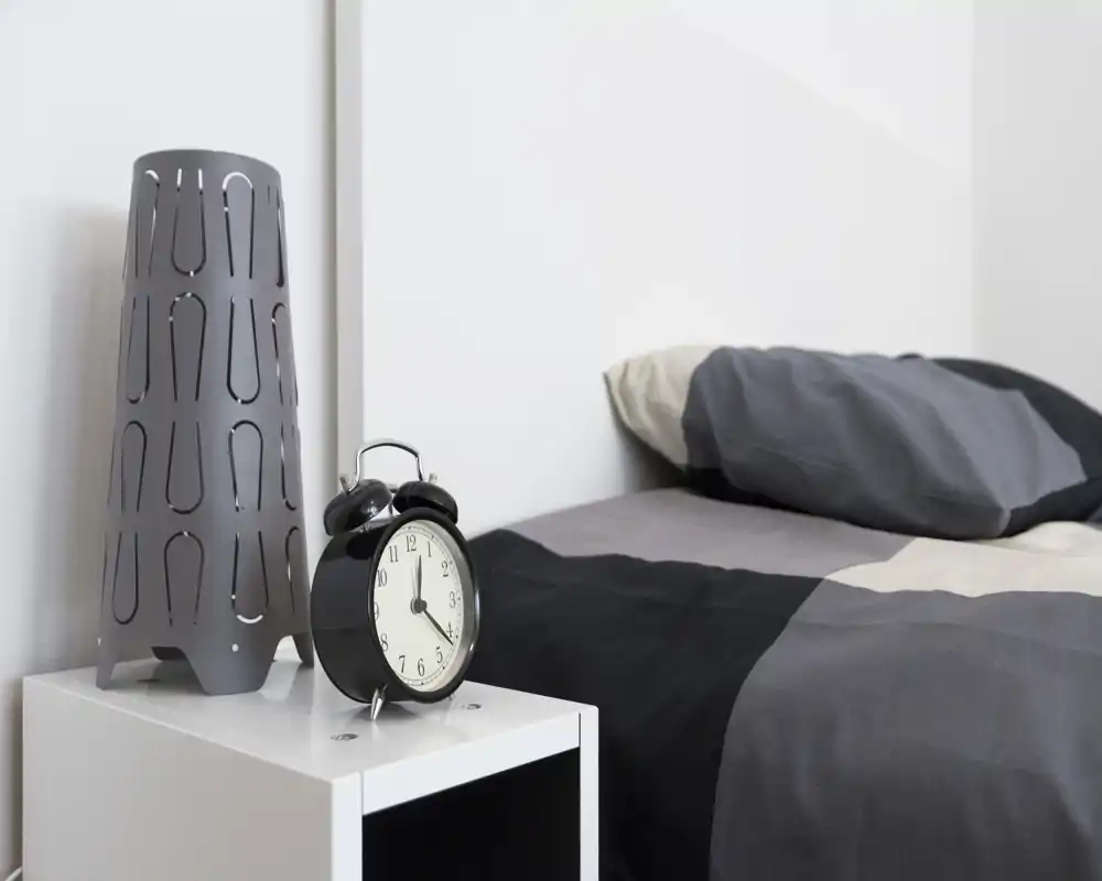 Angolo di camera da letto moderno e minimal: letto rifatto con cuscino, comodino bianco con una sveglia nera e una lampada decorativa.