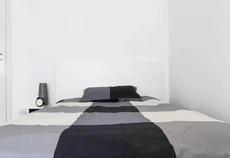 Bright, minimalist bedroom with a centrally placed double bed, monochrome bedding and a bedside table with lamp and clock.