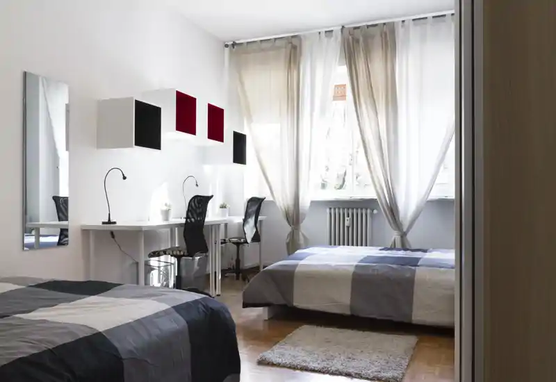 Bright, modern bedroom with two neatly made beds, natural light from a large window, simple desks and soft curtains—ideal for shared rentals or students.