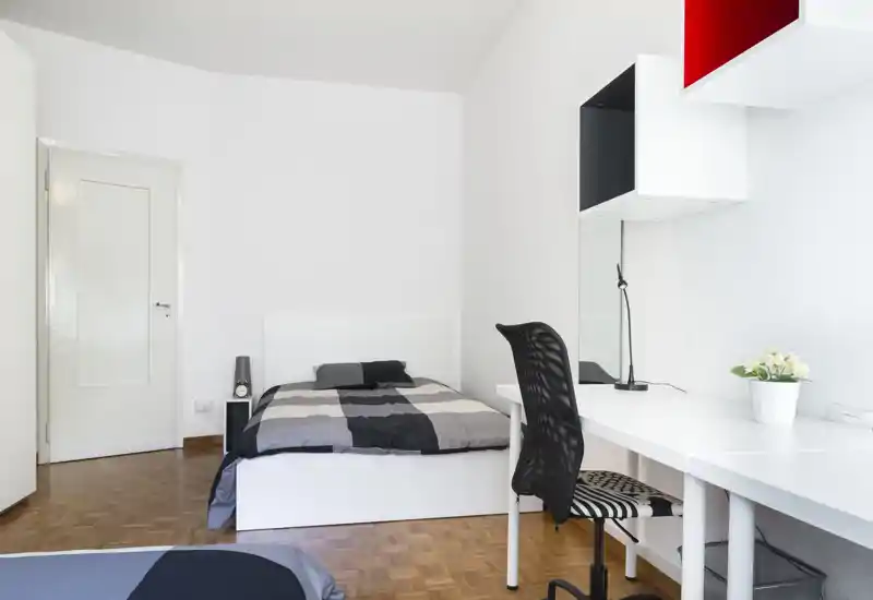 Bright, modern single bedroom with a neatly made bed, a long workspace/desk and shelving. Clean white walls, parquet flooring and simple décor create a calm, contemporary feel ideal for a rental.